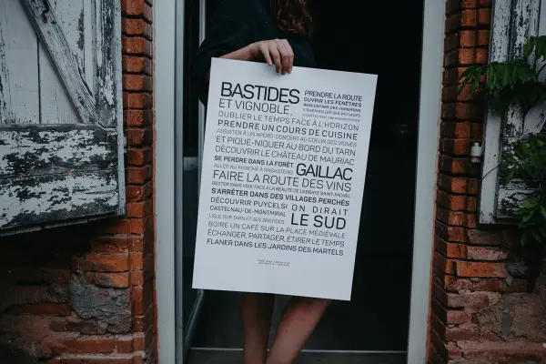 Une femme tenant une pancarte Gaillac, entre bastides et vignoble devant une porte.