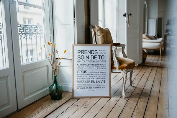 Une pancarte en bois Prends soin de toi posée au sol devant une fenêtre.