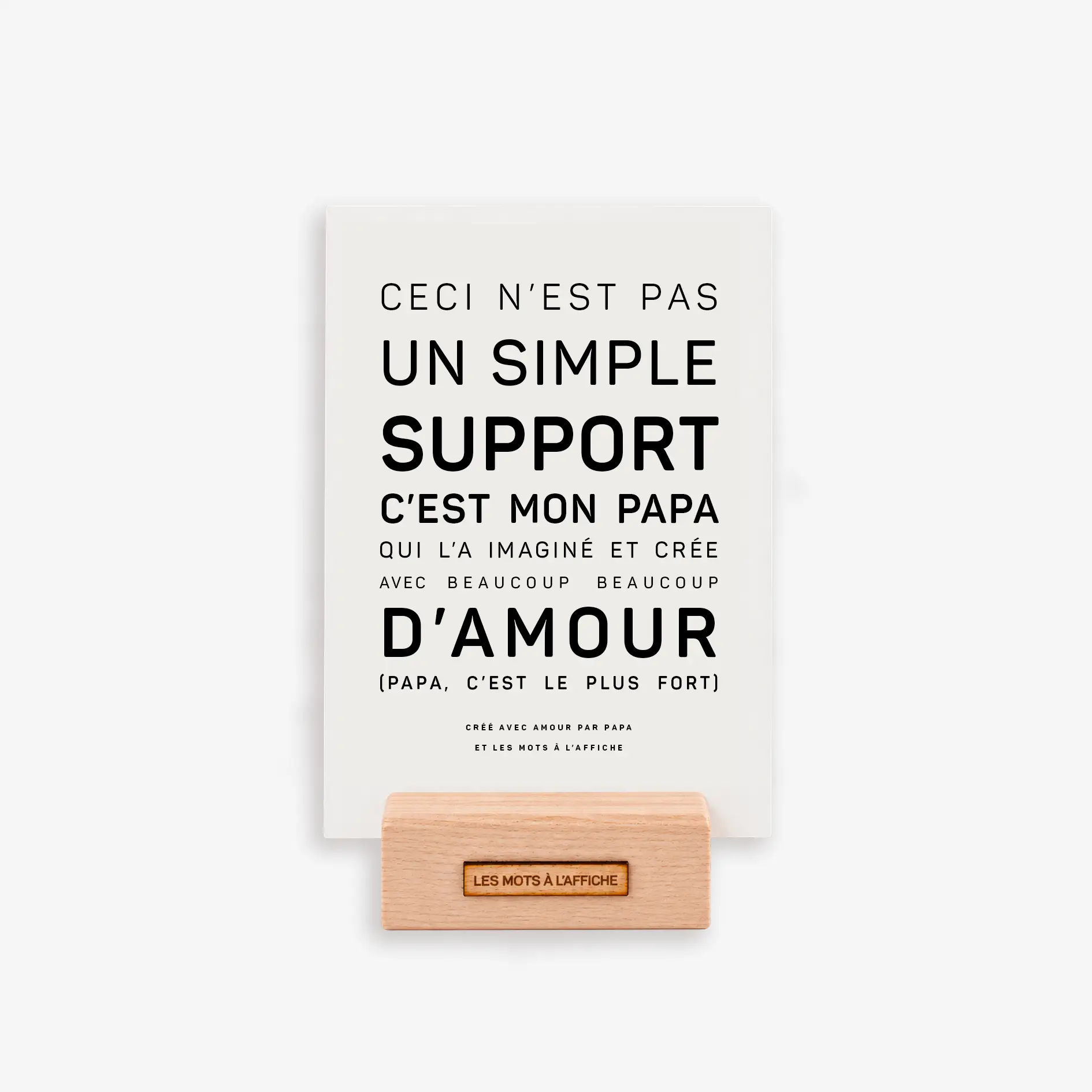 Une carte blanche portant un texte en français sur l'amour d'un père est exposée à la verticale dans un support « Support en bois - mini » portant la mention « Les Mots à l'Affiche », sur un fond blanc uni.
