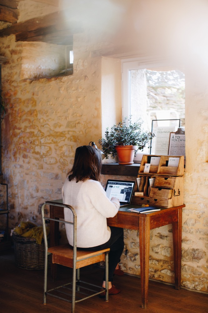 Une femme assise à un bureau avec un ordinateur portable devant une fenêtre, à la recherche de l'idée cadeau parfaite.