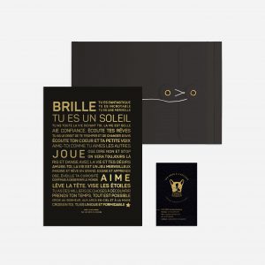 Une enveloppe noire et dorée avec le mot "brille" dessus, contenant un cadeau.