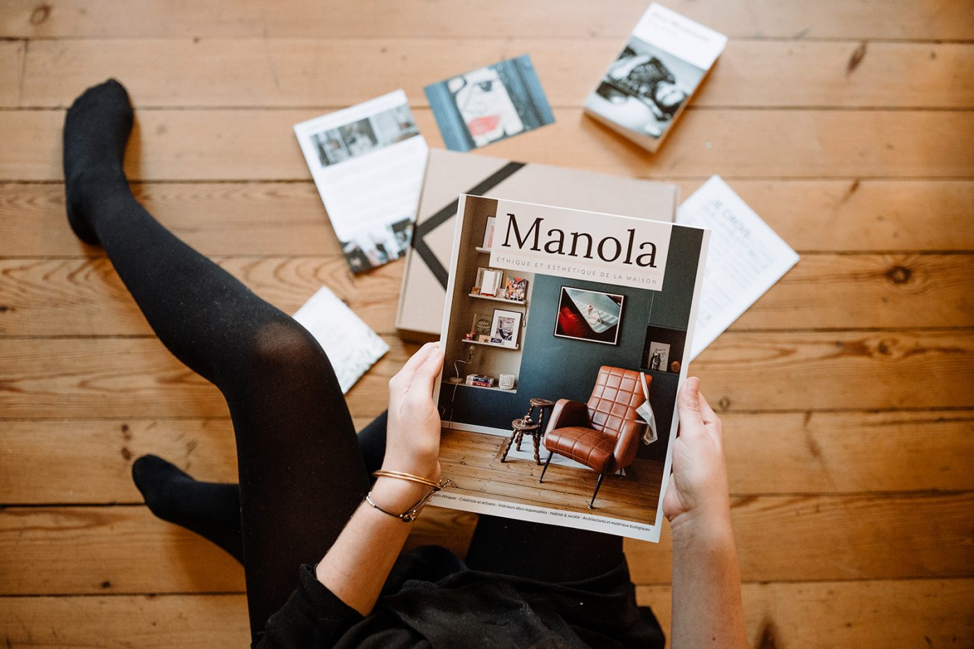Une femme est assise sur un plancher en bois et tient un magazine manola rempli d'idées cadeaux.