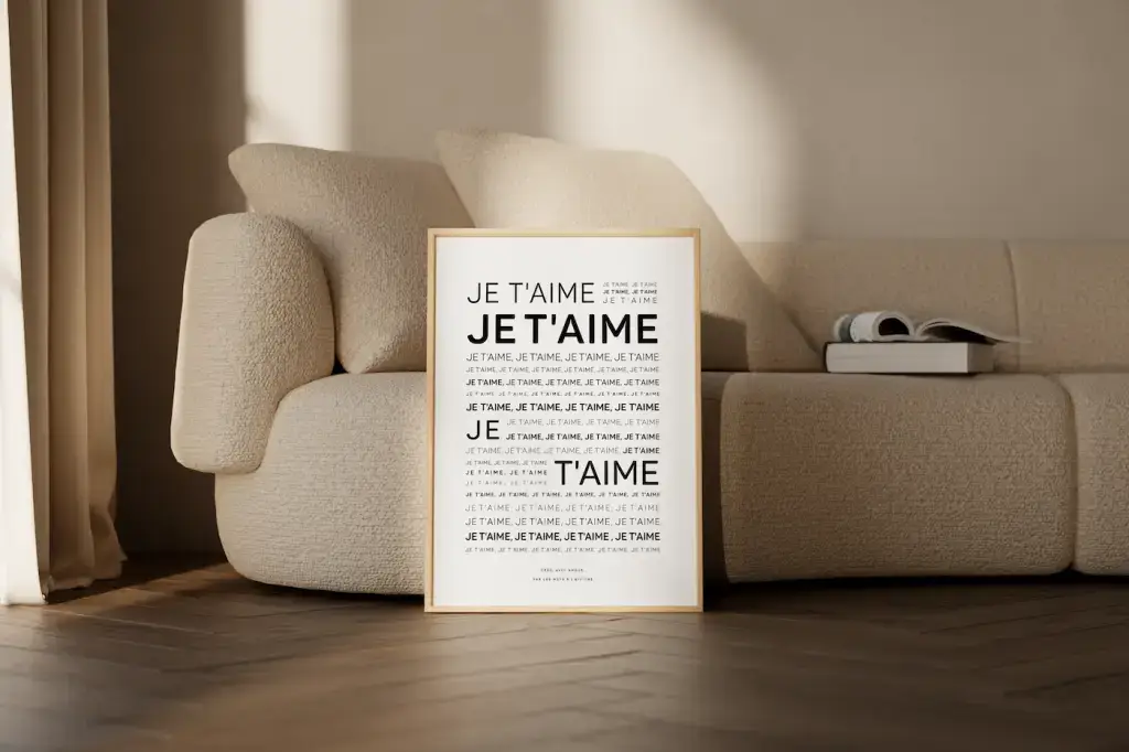 L'affiche encadrée « Mantra : Je t'aime », présentant la phrase dans différents styles, est appuyée contre un canapé beige dans un salon doucement éclairé - une idée cadeau attentionnée pour quelqu'un de spécial, avec un livre sur le canapé.