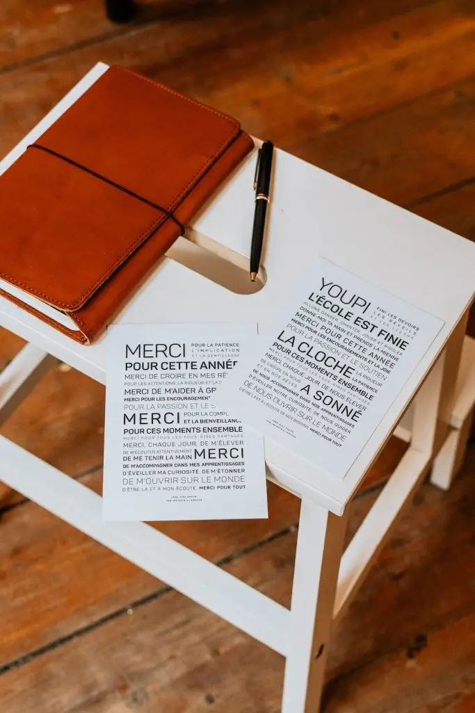 Un cahier marron "Ressources humaines", un stylo noir et deux cartes imprimées en français parfait comme une idée cadeau sont posés sur un tabouret en bois blanc sur un parquet.
