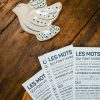 Une Marque-page Les mots, composée de plusieurs cartes imprimées avec des textes en français, est présentée sur une surface en bois à côté d'une figurine d'oiseau en céramique.