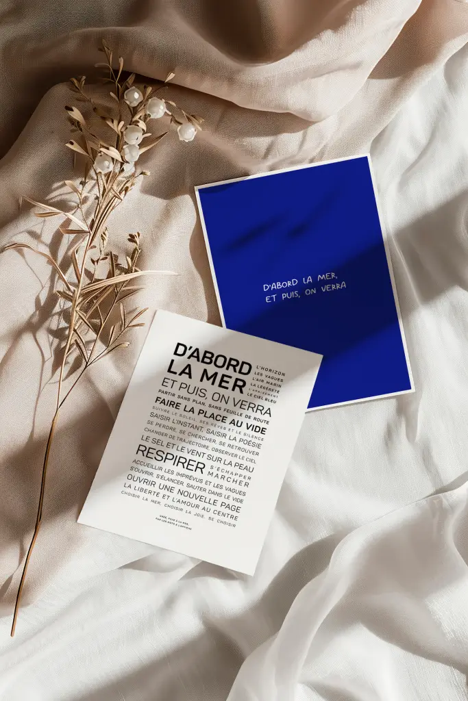 Un brin de fleur séché repose à côté de deux cartes « D'abord, la mer » sur du tissu beige, l'une bleue avec du texte français blanc, l'autre blanche avec du texte français noir en gras.