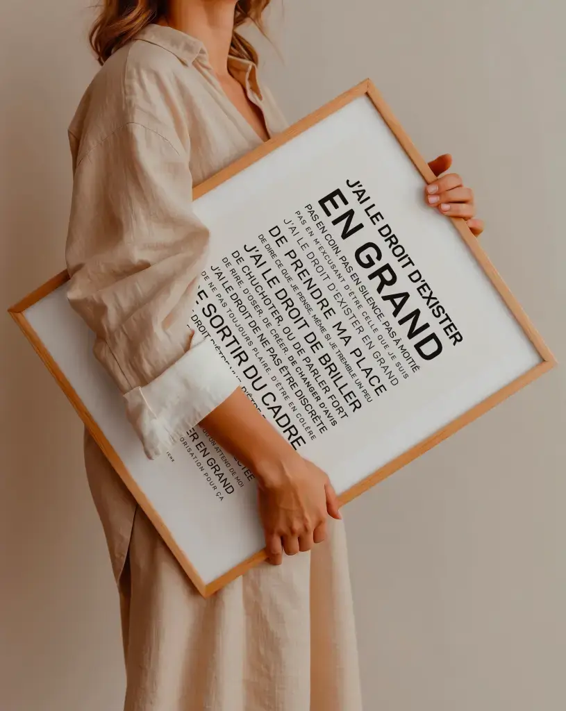 Une personne en tenue beige tient la reproduction encadrée en bois "Tu mérites un amour" avec un texte en français et "EN GRAND" en caractères gras.