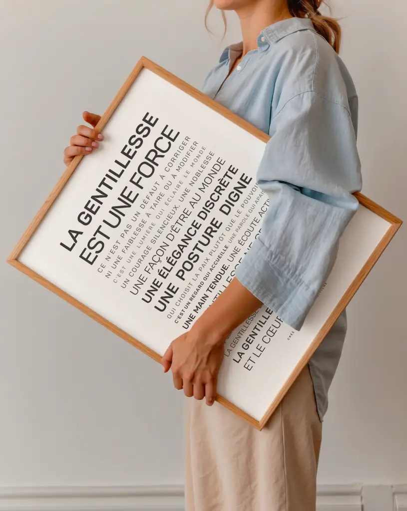 Une personne vêtue d'une chemise bleu clair tient la reproduction encadrée en bois "La gentillesse est une force" avec un texte en français sur un fond uni.