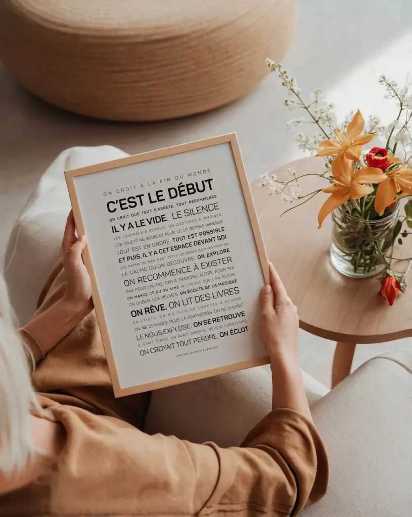 Une personne est assise sur une chaise beige près d'une table avec une composition florale orange, tenant le texte encadré en français "Tu mérites un amour".