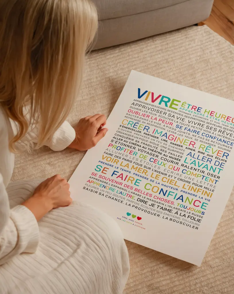 Une personne vêtue de blanc est assise sur un tapis beige, tenant une affiche de motivation française colorée avec diverses phrases en caractères gras et multicolores.