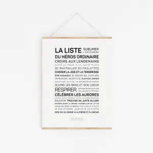 La liste du héros ordinaire" est une affiche blanche avec un texte en français dans différentes tailles de police, qui contient des phrases inspirantes sur les héros ordinaires, la gentillesse, la tendresse et l'amour. Elle est accrochée au mur par une fine ficelle.