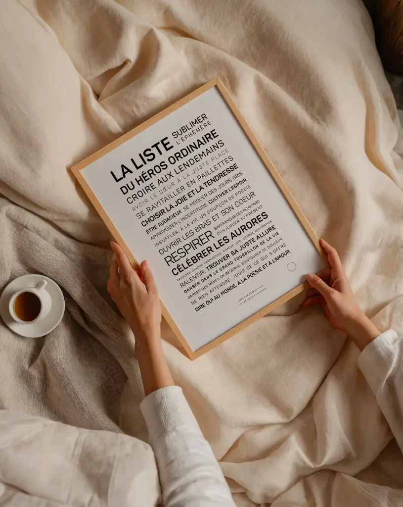 Une personne est assise sur un lit et tient "La liste du héros ordinaire", une liste de citations de motivation encadrée en français, tandis qu'une tasse de café repose à proximité sur la couverture.