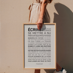 Une femme vêtue d'une robe beige et de baskets se tient sur un trottoir en pierre, tenant une affiche encadrée "Ecrire, se mettre à nu" avec un texte en français contre un mur beige.