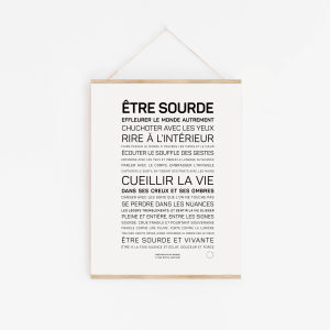 Etre sourde" - une affiche minimaliste avec un texte en français sur l'expérience des sourds, en caractères gras et réguliers, présentée dans un simple cadre en bois avec une ficelle contre un mur blanc.