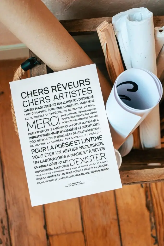 Une lettre en français imprimée, adressée aux artistes et aux rêveurs, repose sur une surface en bois, à côté de papiers enroulés et d'une caisse en bois, vue d'en haut.