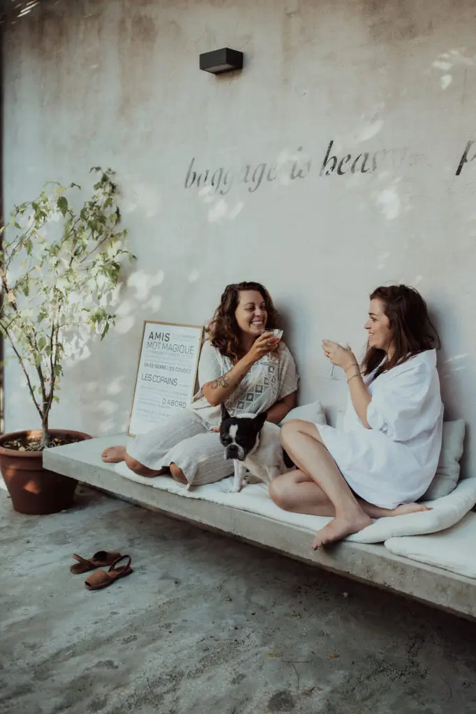 Deux femmes sont assises sur un banc en plein air ; elles rient, un verre à la main, et un petit chien est couché entre elles. Une plante en pot et une pancarte encadrée se trouvent à proximité, et des sandales sont posées par terre.