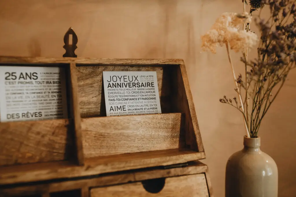Un range-objets en bois contenant des cartes imprimées en français, dont l'une porte l'inscription « Joyeux Anniversaire ». Un vase en céramique avec des fleurs séchées est posé à droite.