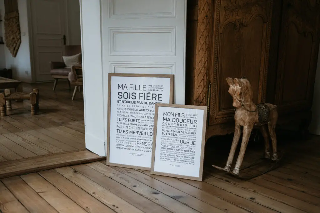 Deux gravures encadrées comportant du texte en français sont posées contre une porte blanche, sur un parquet, près d'un cheval à bascule en bois et d'un meuble en bois richement décoré, dans une pièce chaleureuse.