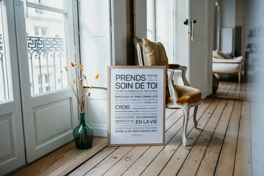 Une citation en français encadrée est posée contre une chaise, sur un parquet, à côté d'un vase contenant des fleurs jaunes, dans une pièce lumineuse et moderne dotée de grandes fenêtres.