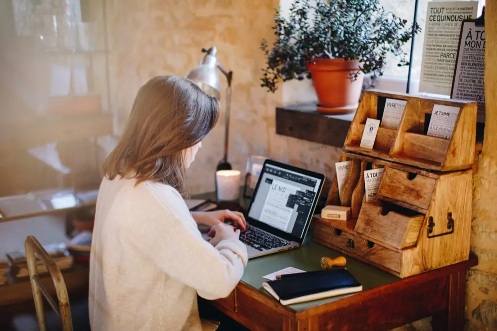 Une personne tape sur un ordinateur portable, assise à un bureau en bois où se trouvent des articles de papeterie, une plante en pot et des feuilles de papier, dans une pièce chaleureuse et tamisée.