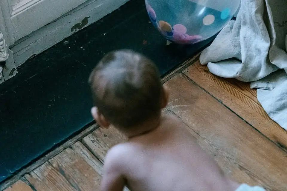 Un bébé en couche rampe sur un parquet, près d'un ballon à pois dégonflé et d'un morceau de tissu froissé.