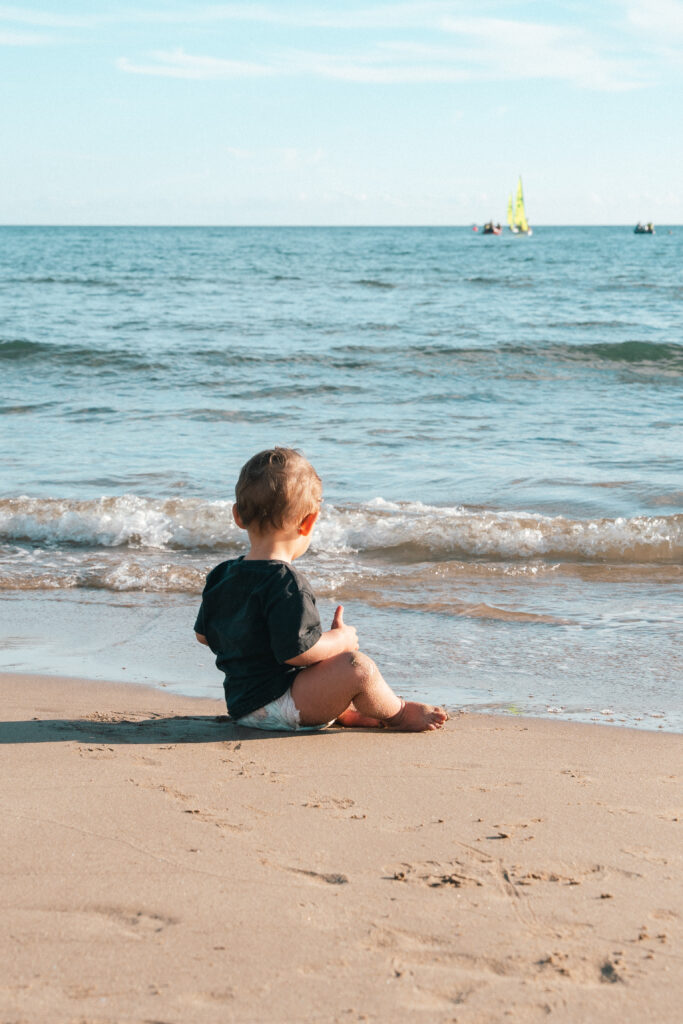 Un petit enfant est assis sur la plage de sable, face à l'océan, avec de douces vagues en arrière-plan par une journée ensoleillée.