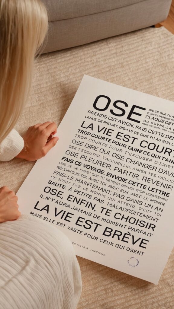 Une personne aux longs cheveux blonds est assise sur un tapis beige, tenant une grande affiche blanche sur laquelle est imprimé en noir un texte de motivation en français en caractères gras.