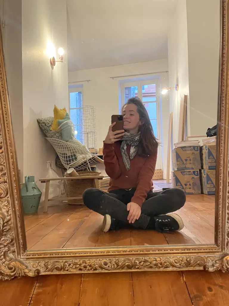 Une femme est assise en tailleur sur un plancher en bois et prend un selfie dans un miroir, dans une pièce où se trouvent des cartons, une chaise et divers objets ménagers.