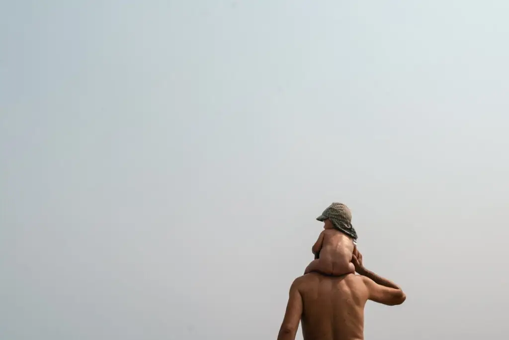 Un adulte torse nu porte sur ses épaules un petit enfant coiffé d'un chapeau ; tous deux sont de dos, face à un ciel clair et pâle.