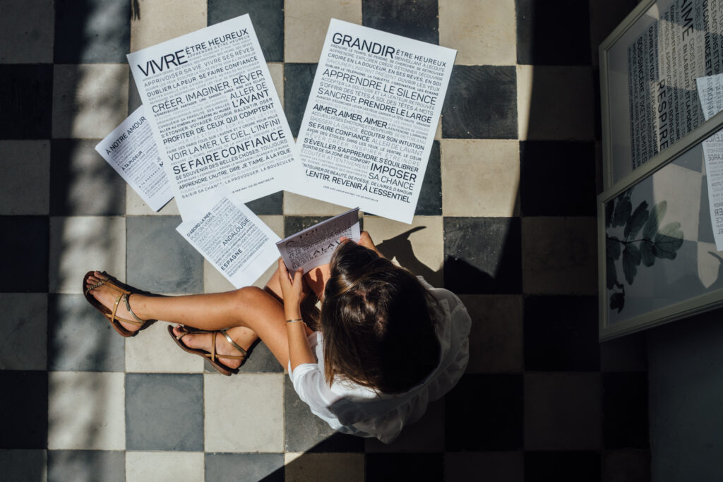 Une femme est assise sur un sol à carreaux et lit un cahier, entourée de grandes feuilles imprimées sur lesquelles figurent des phrases de motivation en français.