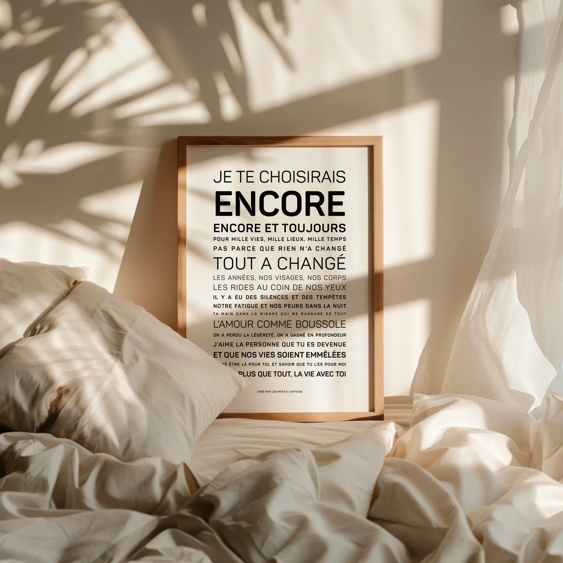 Une reproduction encadrée d'une citation en français repose sur un lit défait, garni de draps blancs, baigné par la lumière chaude du soleil et les ombres douces projetées par les plantes voisines.