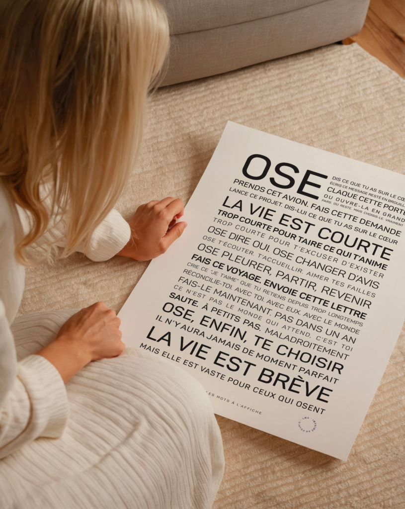 Une femme vêtue d'une robe blanche est assise sur un tapis beige ; elle tient dans ses mains une affiche française motivante qui invite à profiter pleinement de la vie et à prendre des risques, et la regarde attentivement.