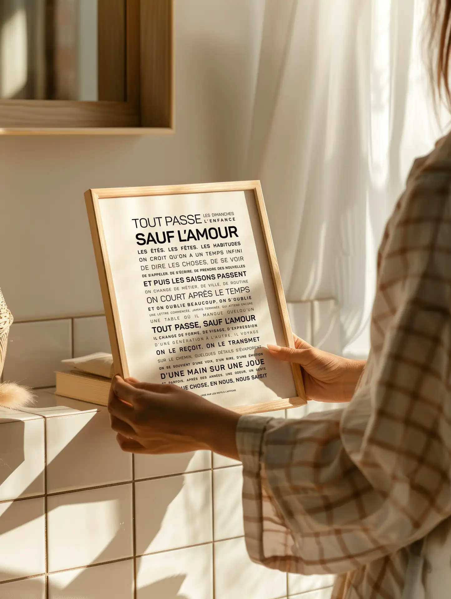 Une personne vêtue d'une chemise à carreaux tient, à la verticale, une reproduction encadrée d'un texte en français sur le plan de travail carrelé d'une salle de bains, tandis que la lumière du soleil pénètre à flots par la fenêtre.