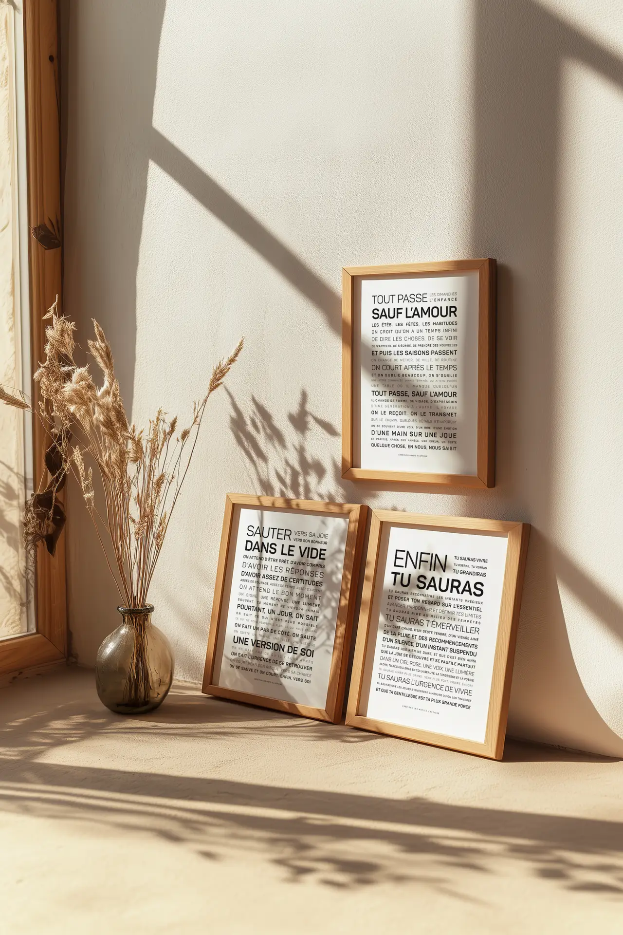 Trois gravures encadrées comportant du texte en français sont exposées sur une surface en bois clair, près d'un vase contenant des herbes séchées, le tout se détachant sur un mur baigné de soleil qui projette des ombres douces.