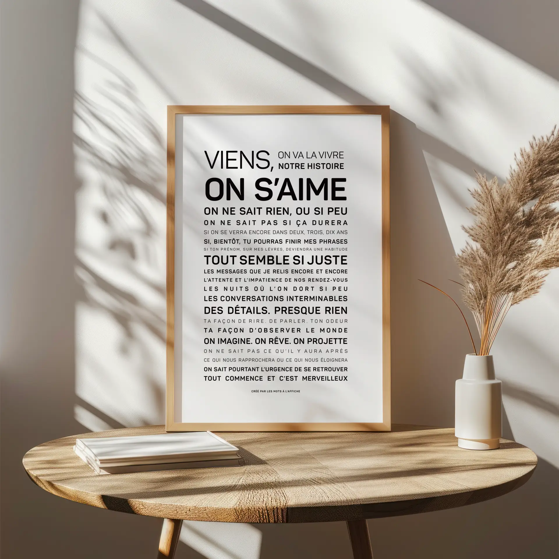 Une affiche encadrée portant un texte en français est posée sur une table en bois, à côté d'un vase contenant de l'herbe séchée, tandis que la lumière du soleil projette des ombres sur le mur blanc situé derrière.