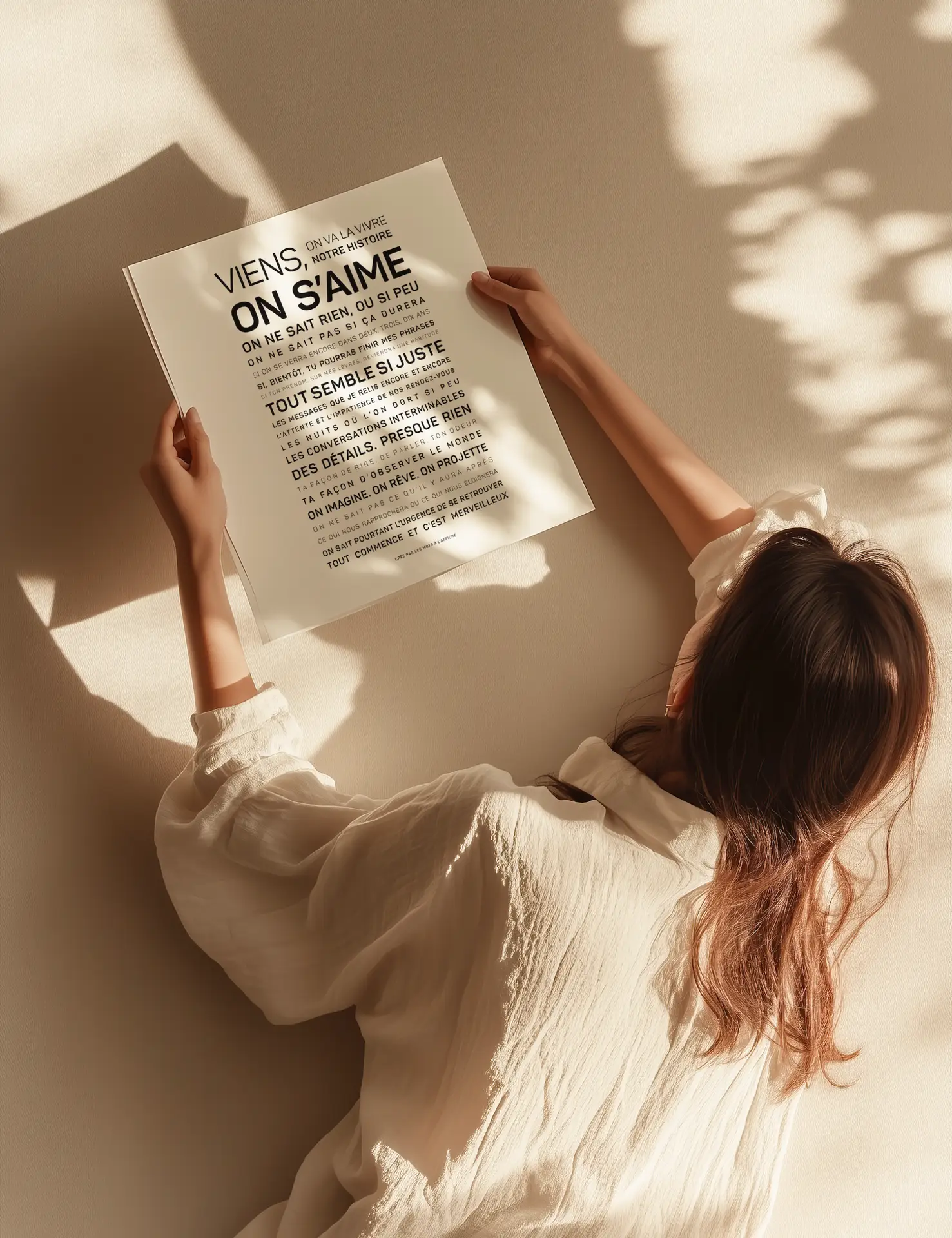 Une femme vêtue d'un chemisier blanc est allongée sur le sol, tenant dans ses mains une estampe carrée sur laquelle figure un texte en français. Les rayons du soleil et les ombres des feuilles se projettent sur elle et sur l'estampe.