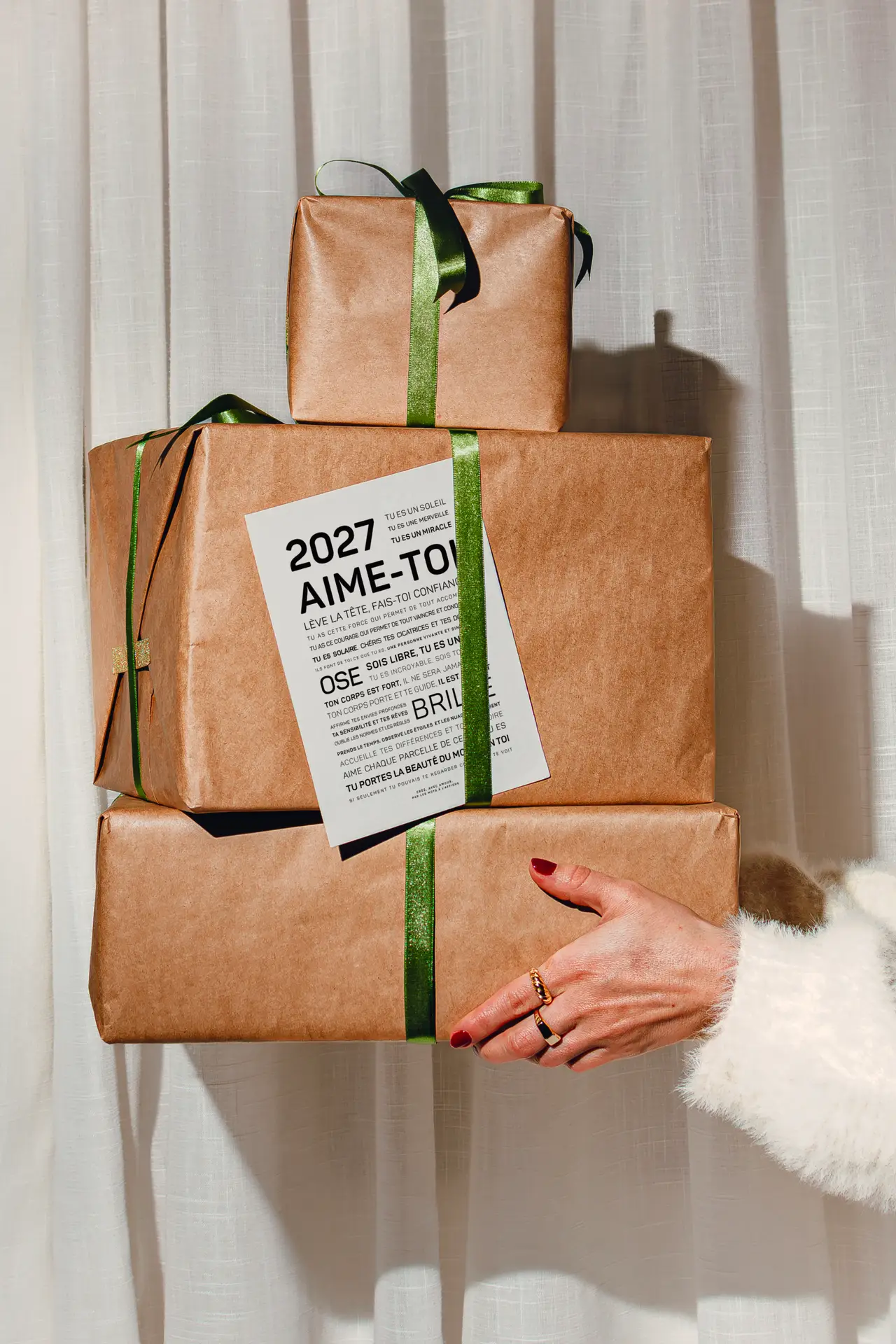 Trois cadeaux emballés dans du papier kraft et ornés de rubans verts sont empilés ; ils sont tenus par une personne vêtue de blanc, et un papier sur le thème de 2027 est fixé sur le cadeau du milieu.