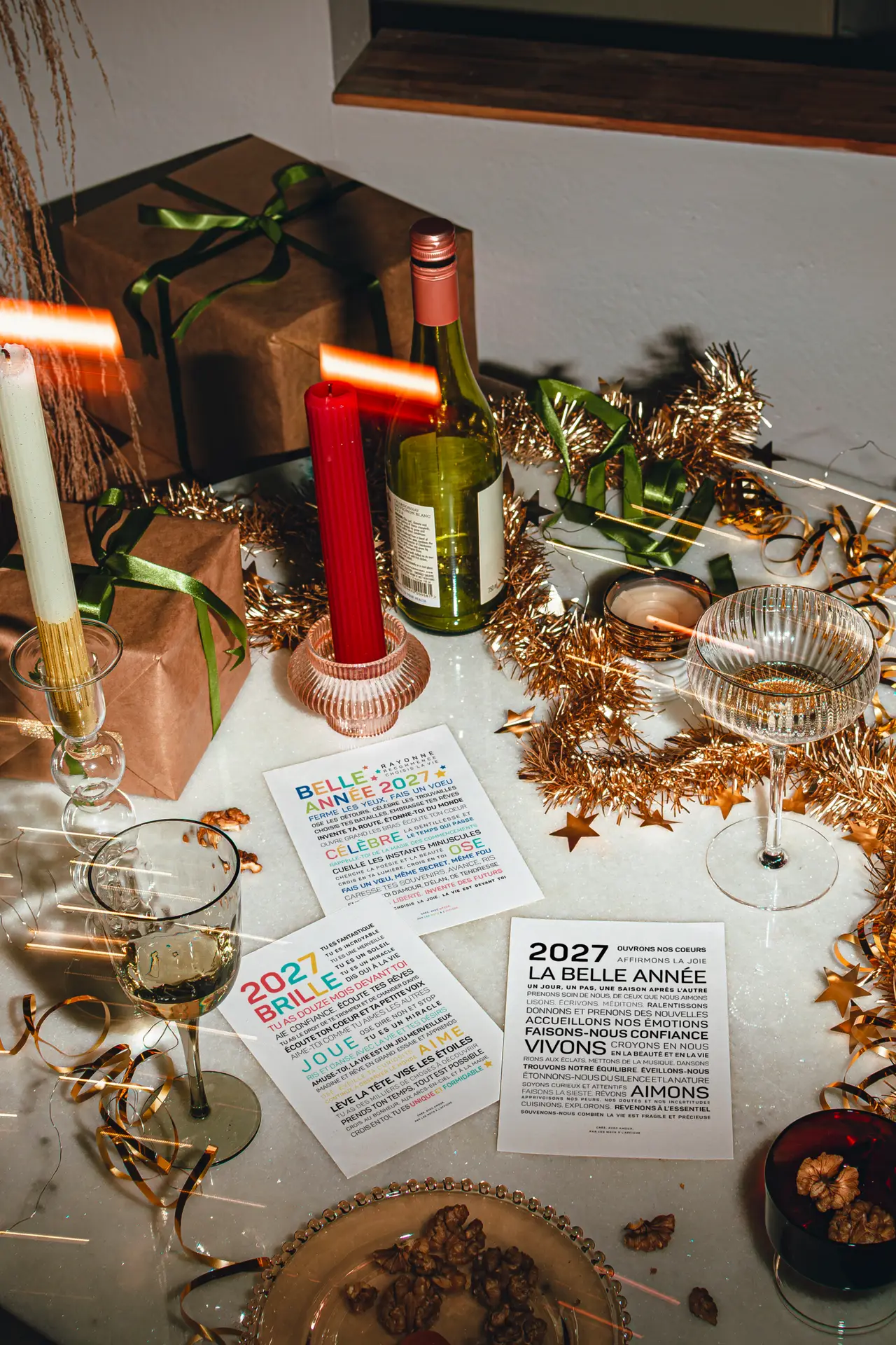 Une table de fête avec des cadeaux emballés, une bouteille de vin, des verres, des bougies, des décorations dorées et trois cartes sur le thème du Nouvel An, le tout posé sur un plateau en marbre.