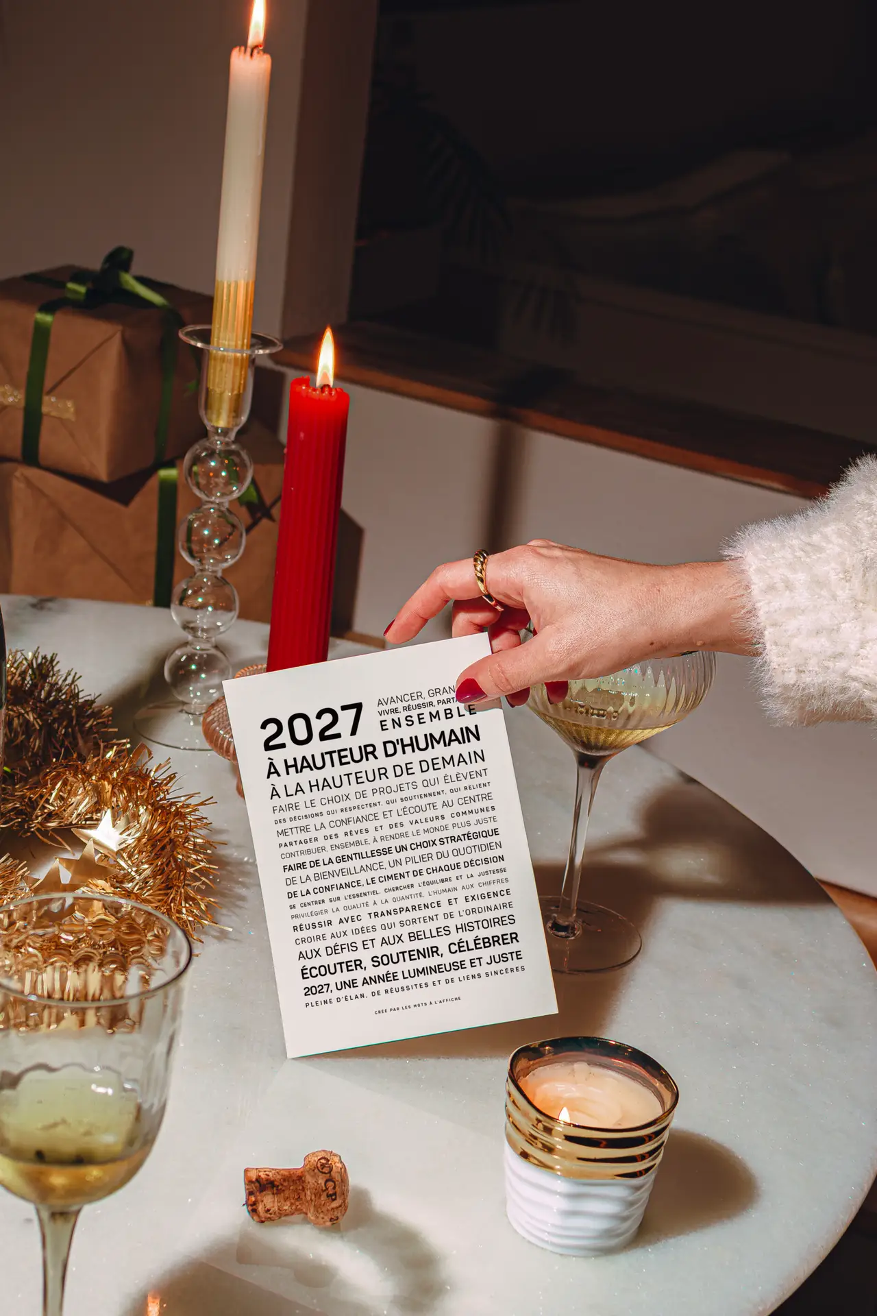 Une main tient une carte sur laquelle figure un texte en français, à côté de bougies, d'un cadeau emballé, de boissons et de décorations de fête, le tout posé sur une table en marbre.