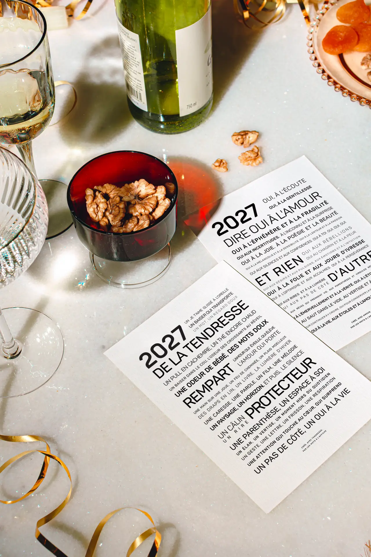 Deux verres, une bouteille de vin verte, des noix et des confettis entourent deux cartes posées sur une table en marbre. Les cartes portent un texte en français en caractères gras, où l'année 2027 est mise en évidence.