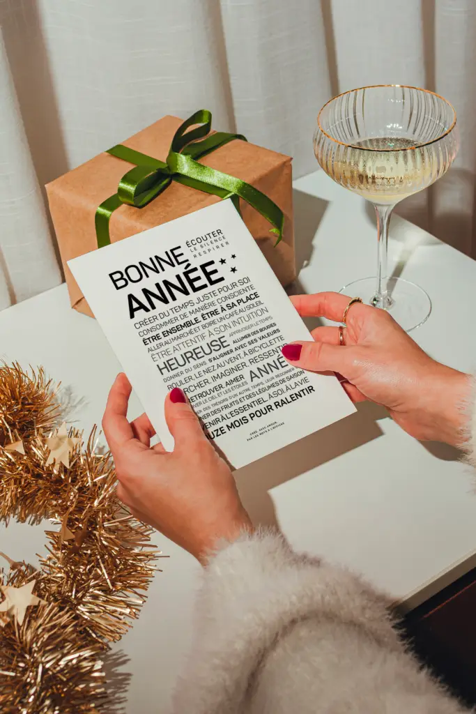 Une personne tient une carte de vœux de Nouvel An en français, à côté d'un cadeau emballé, d'un verre de boisson pétillante et de guirlandes dorées, posés sur une table blanche.