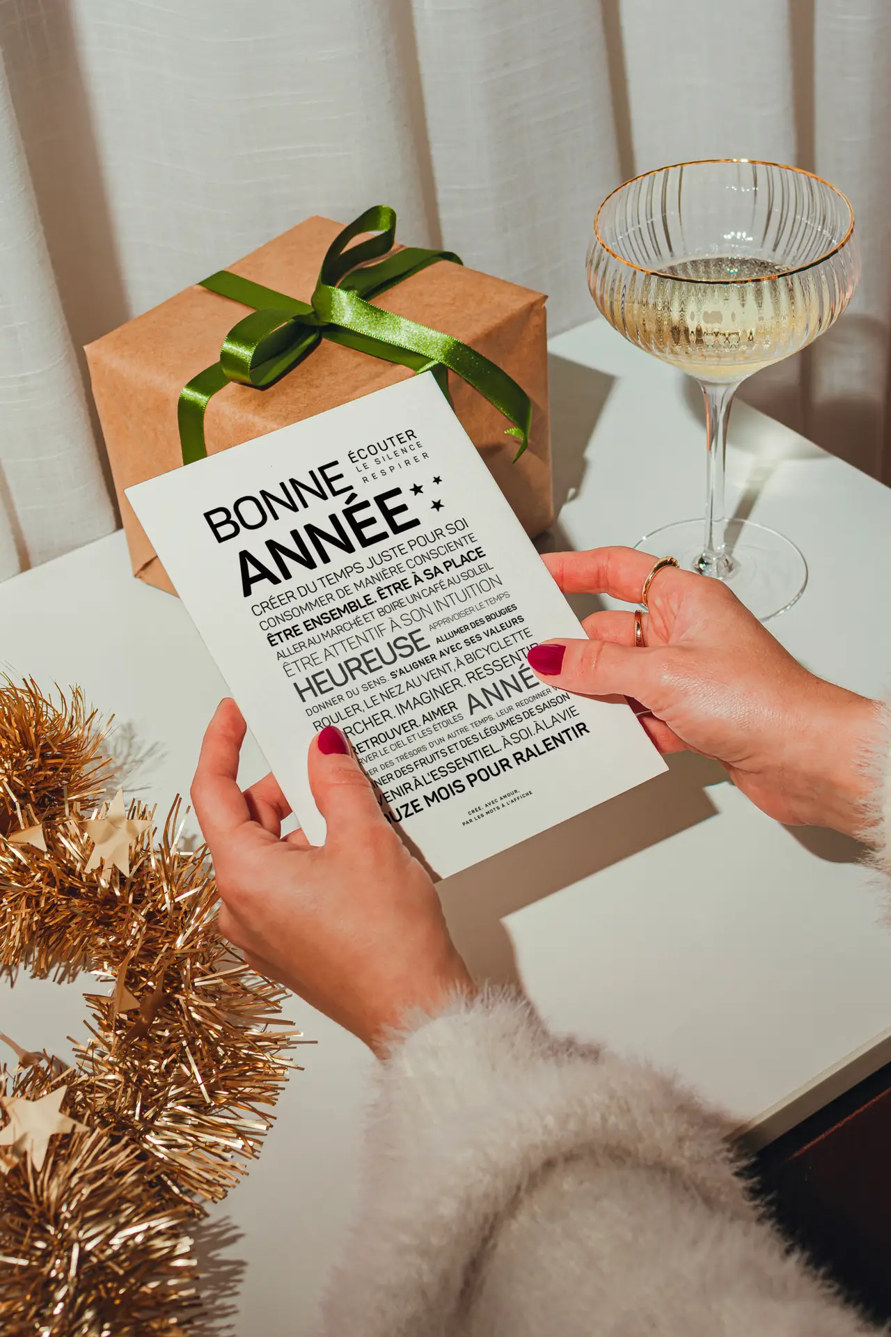 Une personne tient une carte de vœux de Nouvel An en français, à côté d'un cadeau emballé, d'un verre de boisson pétillante et de guirlandes dorées, posés sur une table blanche.