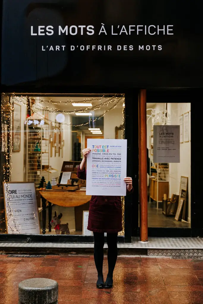 Une personne se tient sur un trottoir mouillé devant un magasin, tenant une grande affiche qui lui cache le visage et le torse. L’enseigne du magasin indique « LES MOTS À L’AFFICHE ».