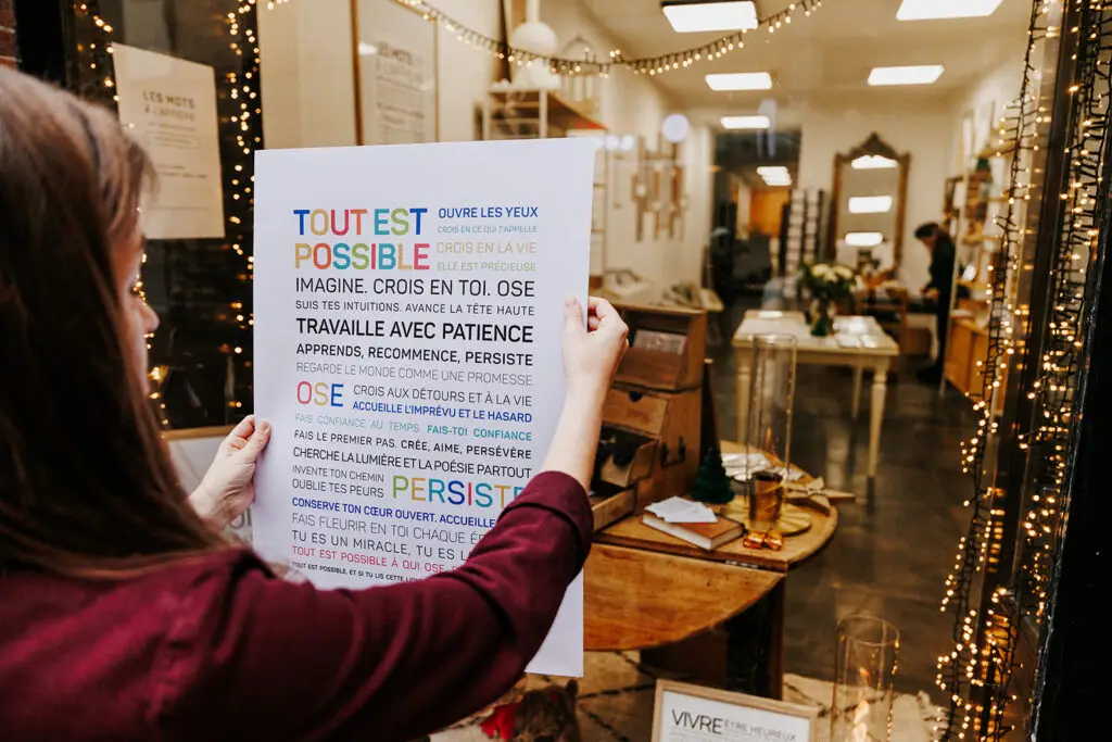 Une personne tient une affiche de motivation française aux couleurs vives à l'intérieur d'une boutique bien éclairée, décorée de guirlandes lumineuses, dont on aperçoit le mobilier et les articles exposés à travers la vitrine.