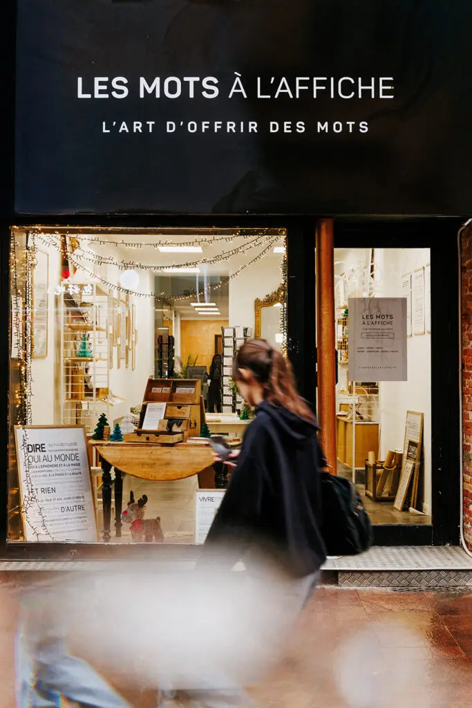 Une personne passe devant une vitrine sur laquelle est accrochée une enseigne indiquant « LES MOTS À L’AFFICHE L’ART D’OFFRIR DES MOTS ». La vitrine présente des œuvres textuelles encadrées et des objets de décoration en bois exposés à l’intérieur de la boutique.