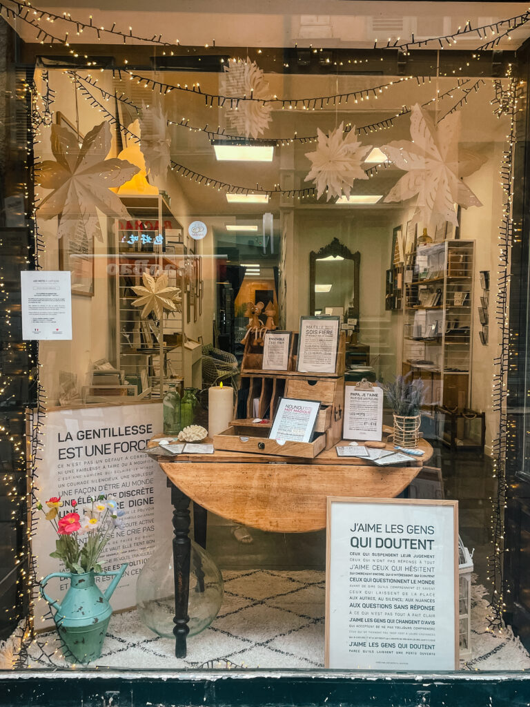 Vitrine comportant un bureau en bois, des citations en français encadrées, des livres, un vase avec des fleurs et de grands flocons de neige en papier blanc, le tout agrémenté de guirlandes lumineuses.